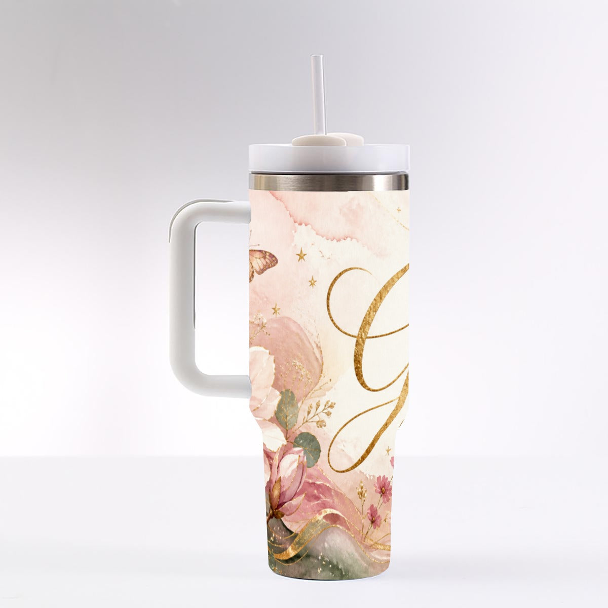 A handled tumbler with a straw, featuring a pastel pink and cream floral design with gold cursive lettering and small gold stars, shown on a light gray studio background.