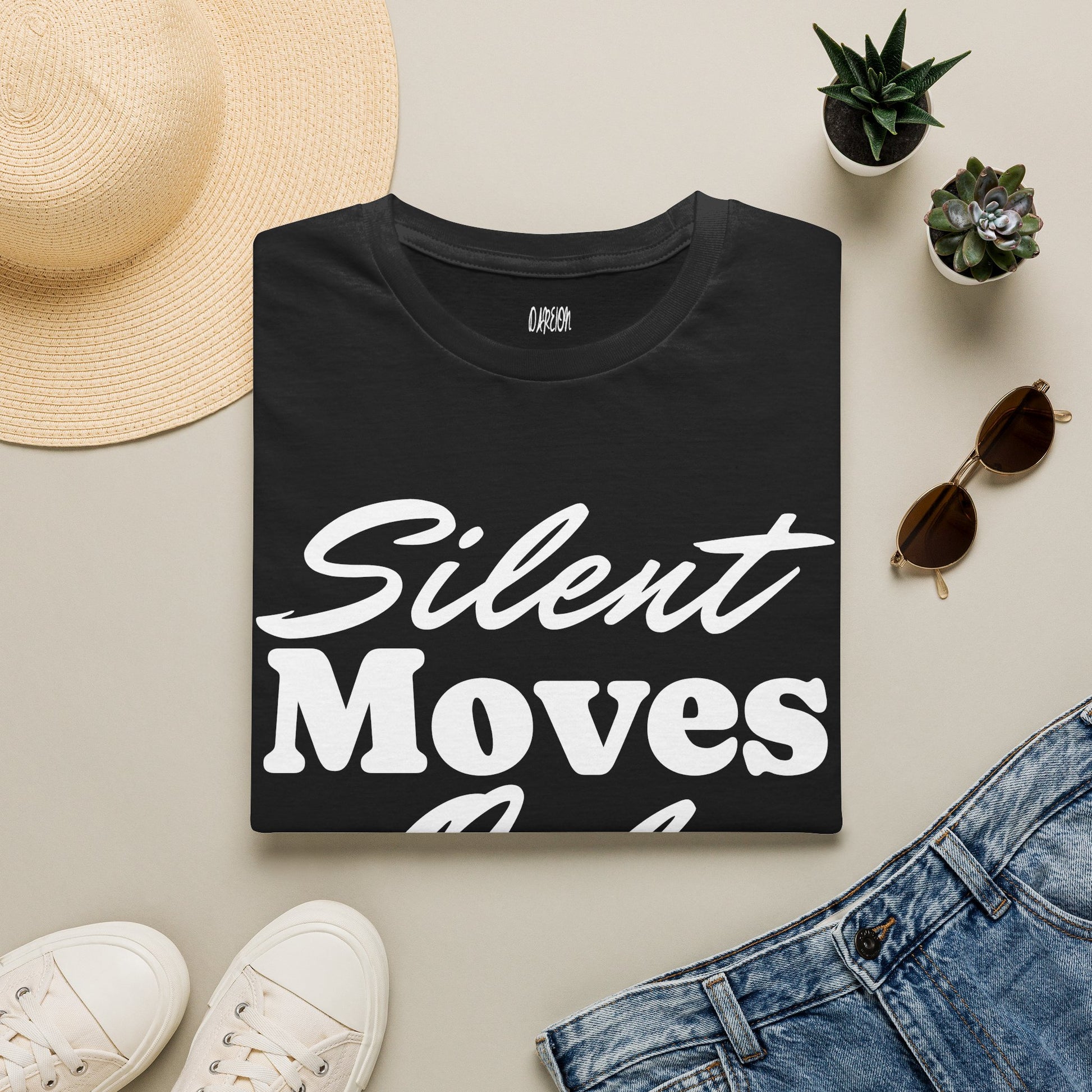 Black t-shirt with 'Silent Moves' text on a light surface with casual items like a hat, sunglasses, and jeans.