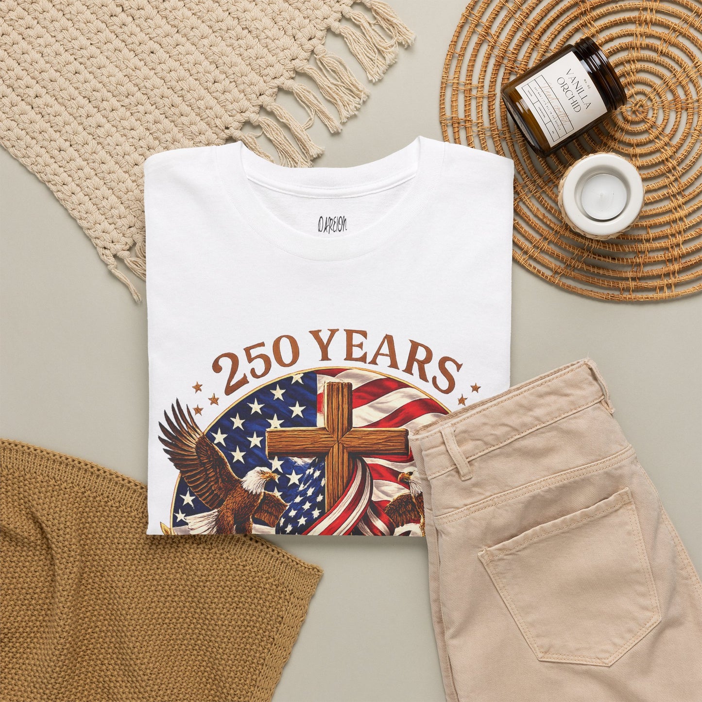 White t-shirt with '250 Years' design featuring an eagle, cross, and American flag on a beige surface with a woven mat and candles.