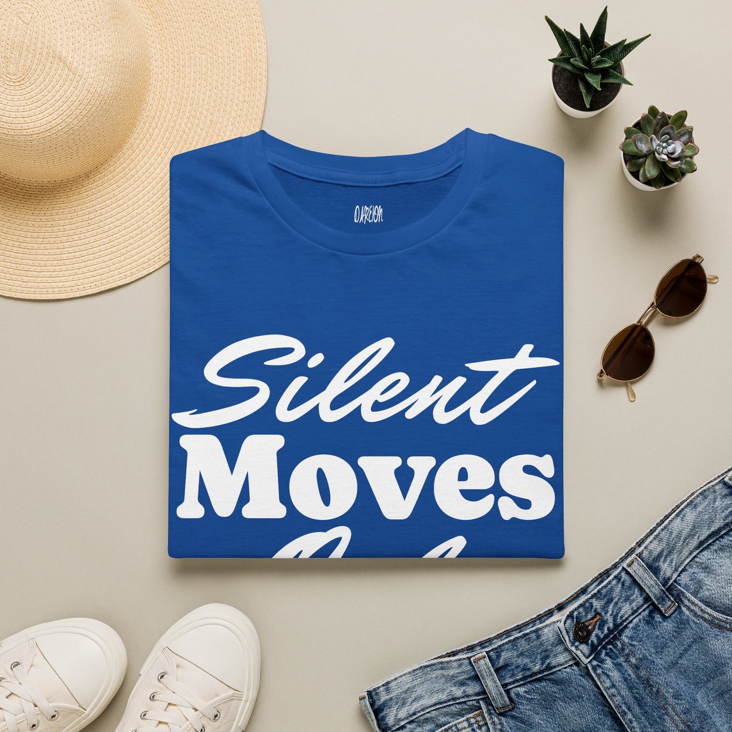 Blue t-shirt with 'Silent Moves' text on a light surface with accessories like a hat, sunglasses, and jeans.
