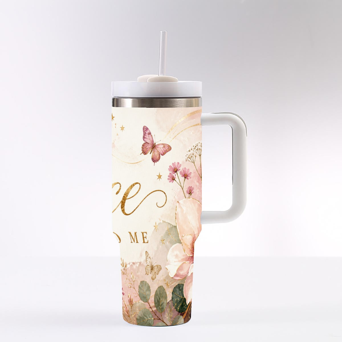 A tall handled tumbler with a white lid and straw, featuring a watercolor-style floral and butterfly print in soft pink, beige, and green tones with gold script lettering, shown on a light gray background.