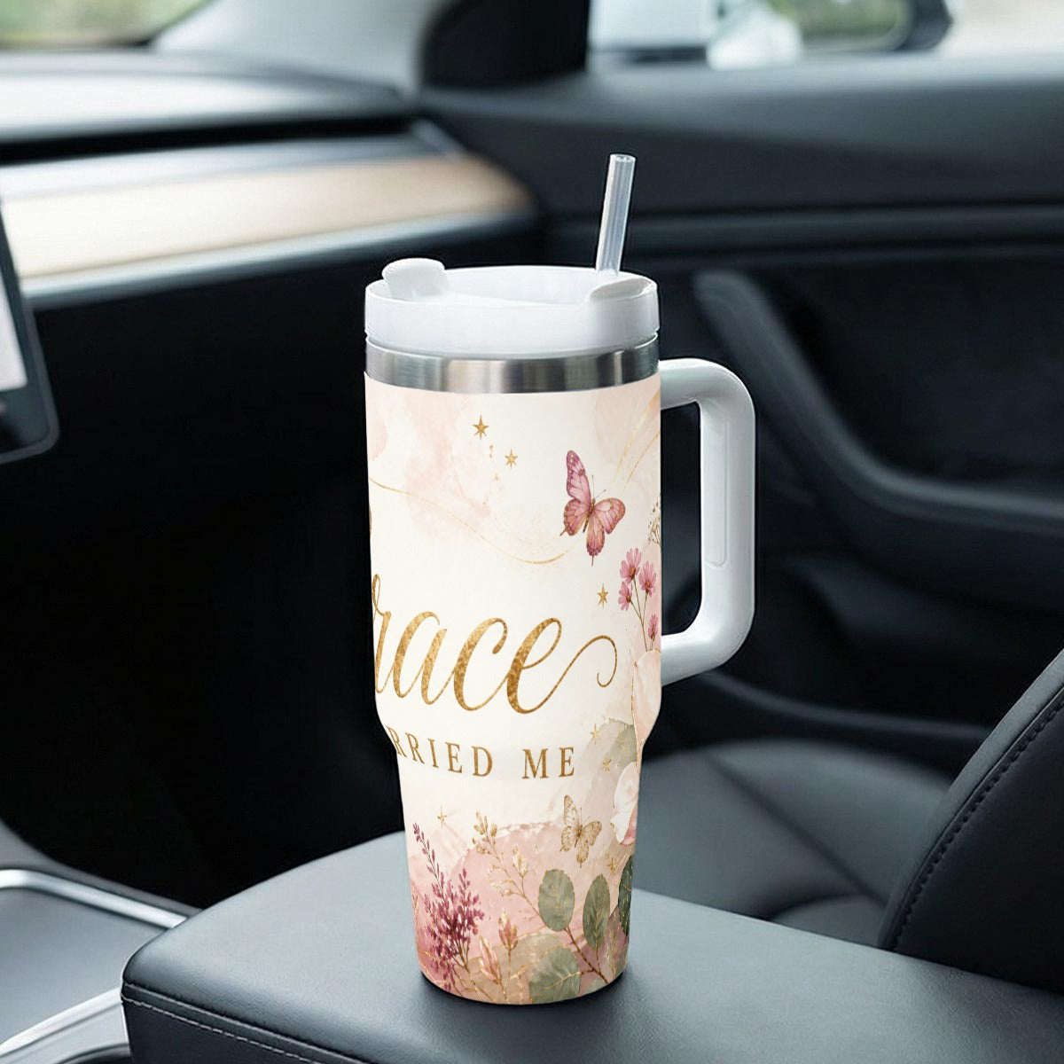 A travel tumbler with handle, a lid and straw, featuring a floral and butterfly design with the text 'Grace Married Me' in gold, resting on a car's center console.