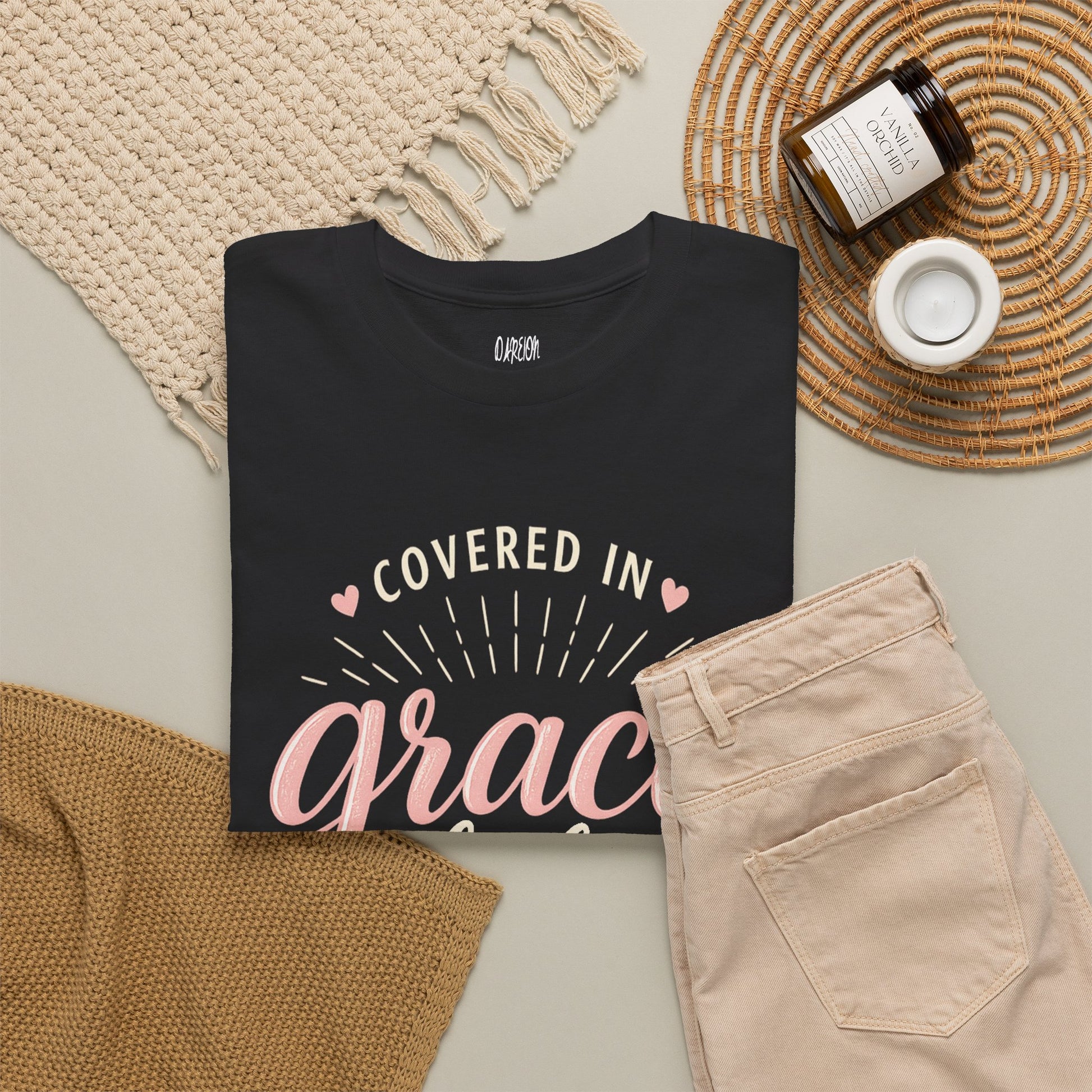 Black t-shirt with 'Covered in Grace' text on a neutral surface with pants and decor items.