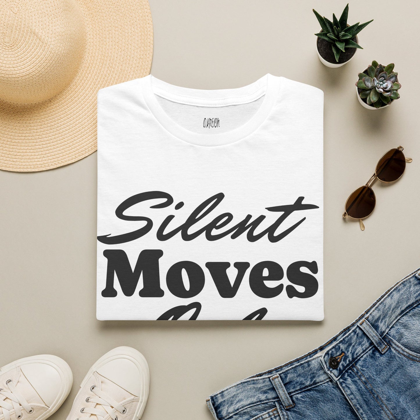 White t-shirt with 'Silent Moves' text, surrounded by casual clothing and accessories on a beige surface.