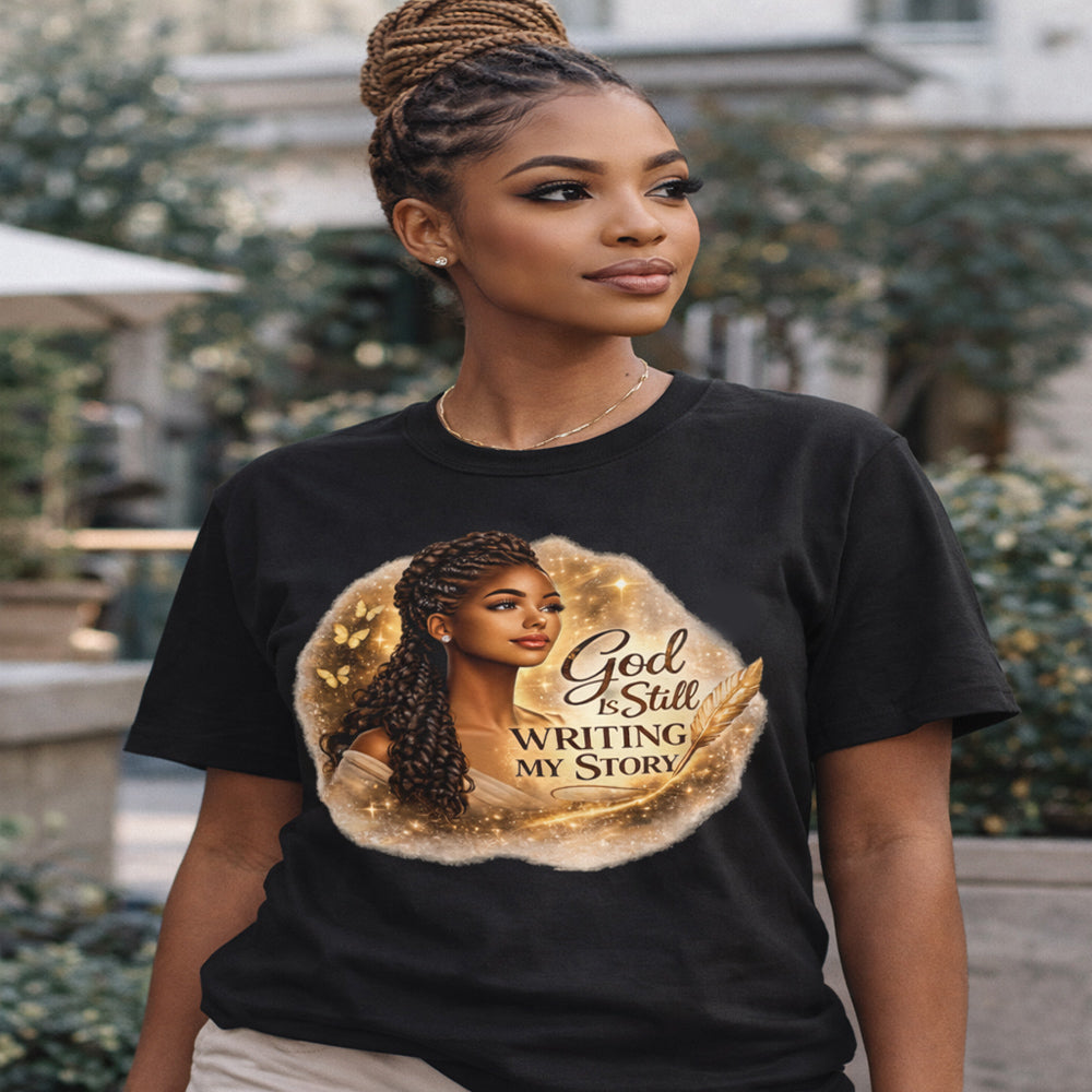 Woman wearing a black t-shirt with a graphic of a woman with braids and text 'God is Still Writing My Story'.